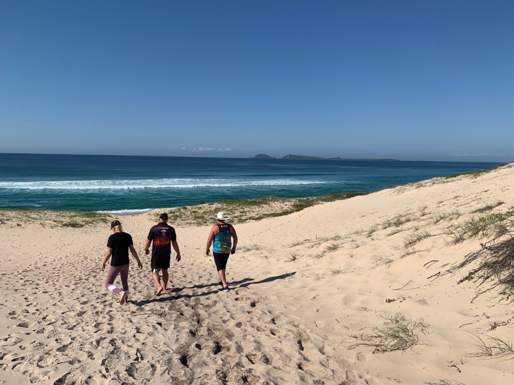 3 people walking across the sand dunes towards the wave break on Mungo Beach. In the distance Broughton Island can be seen.