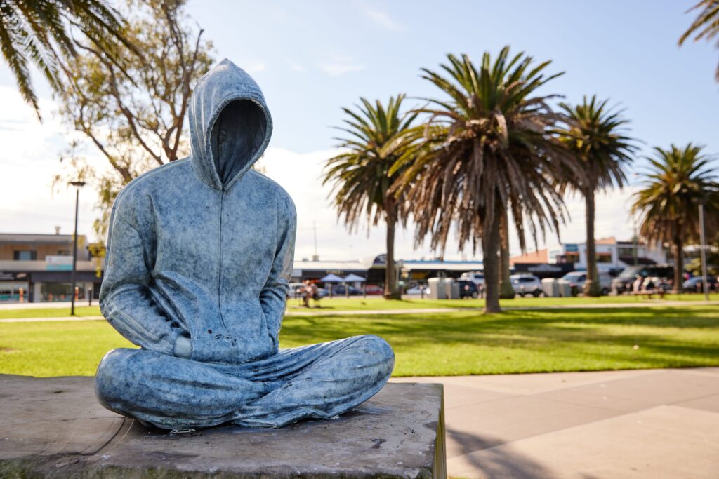 A sculpture on a person sitting on a concrete block in tracksuit and hoodie overlooking Lake Macquarie at Warners Bay foreshore