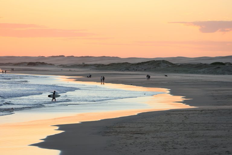 Surfer exits the water and people enjoy a walk along the beach as the sun sets over the Stockton Sand dunes.