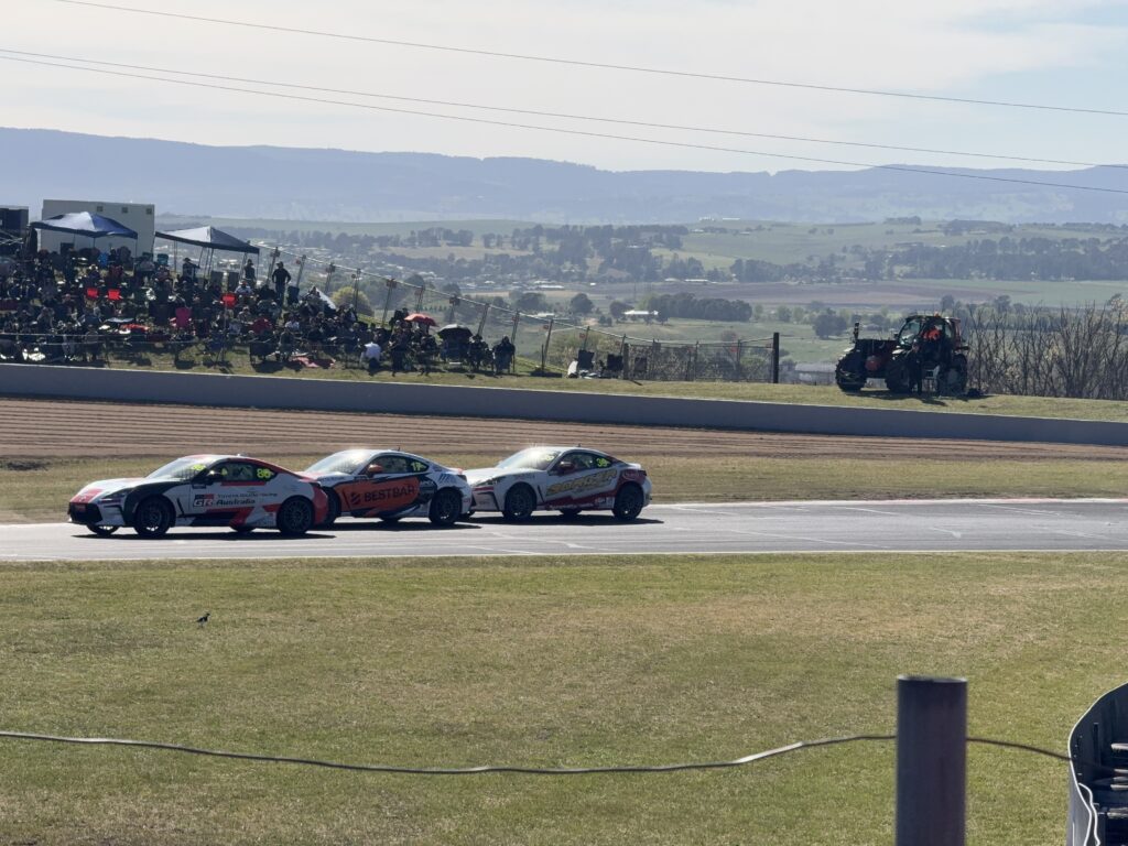 3 Toyota GAZOO cars in bumper to bumper racing at the Chase with crowds watching on the hill and farmlands in the distance