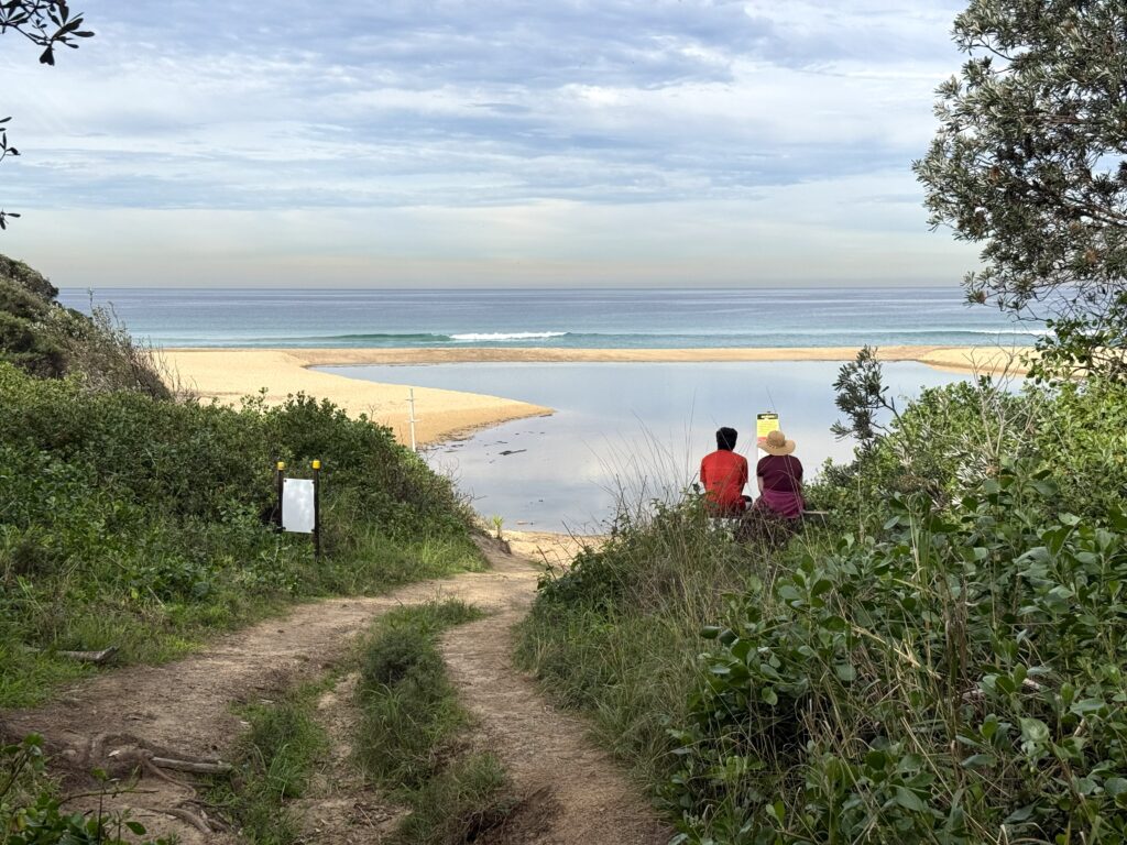 A couple of hikers resting at The Bench after walking the Yuelarbah Walking Track to Glenrock Lagoon with views of Glenrock Beach to the ocean and coal ships on the horizon