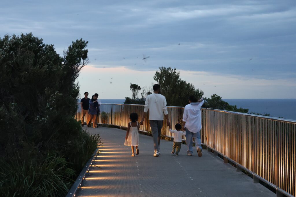 Family walks along the ANZAC Memorial Walk at dusk as lights illuminate the walkway.