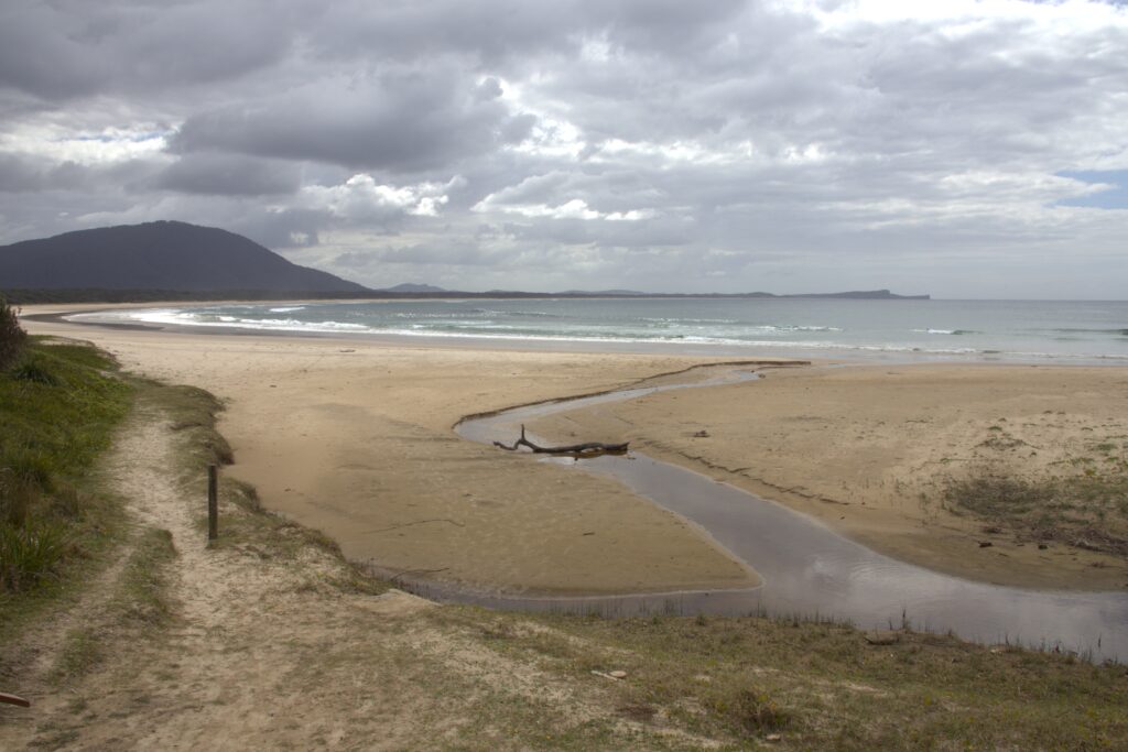 A creek meanders across the beach at Diamond Head Beach with North Brother Mountain in the background on an overcast day.