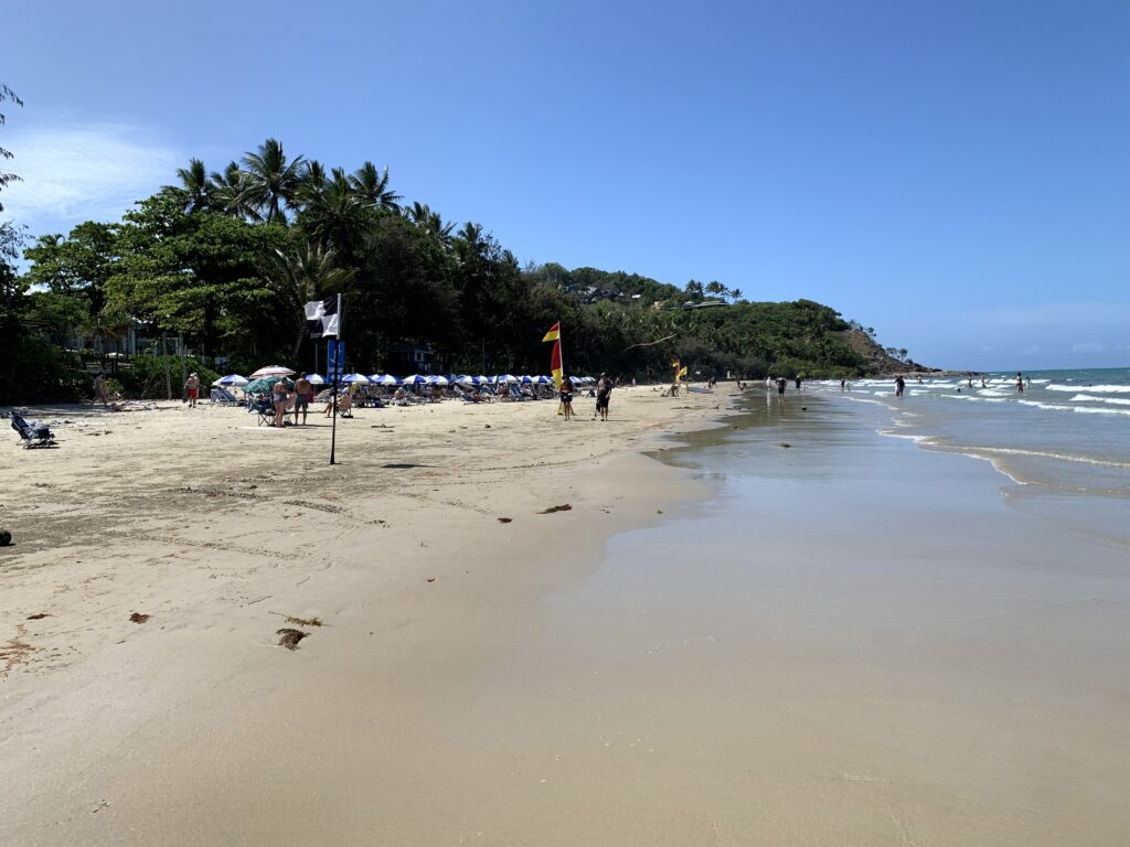 People enjoying the tropical sunshine under blue and white beach umbrellas and swimming in the turquoise waters between the red and yellow flags on Four Mile Beach in Port Douglas.