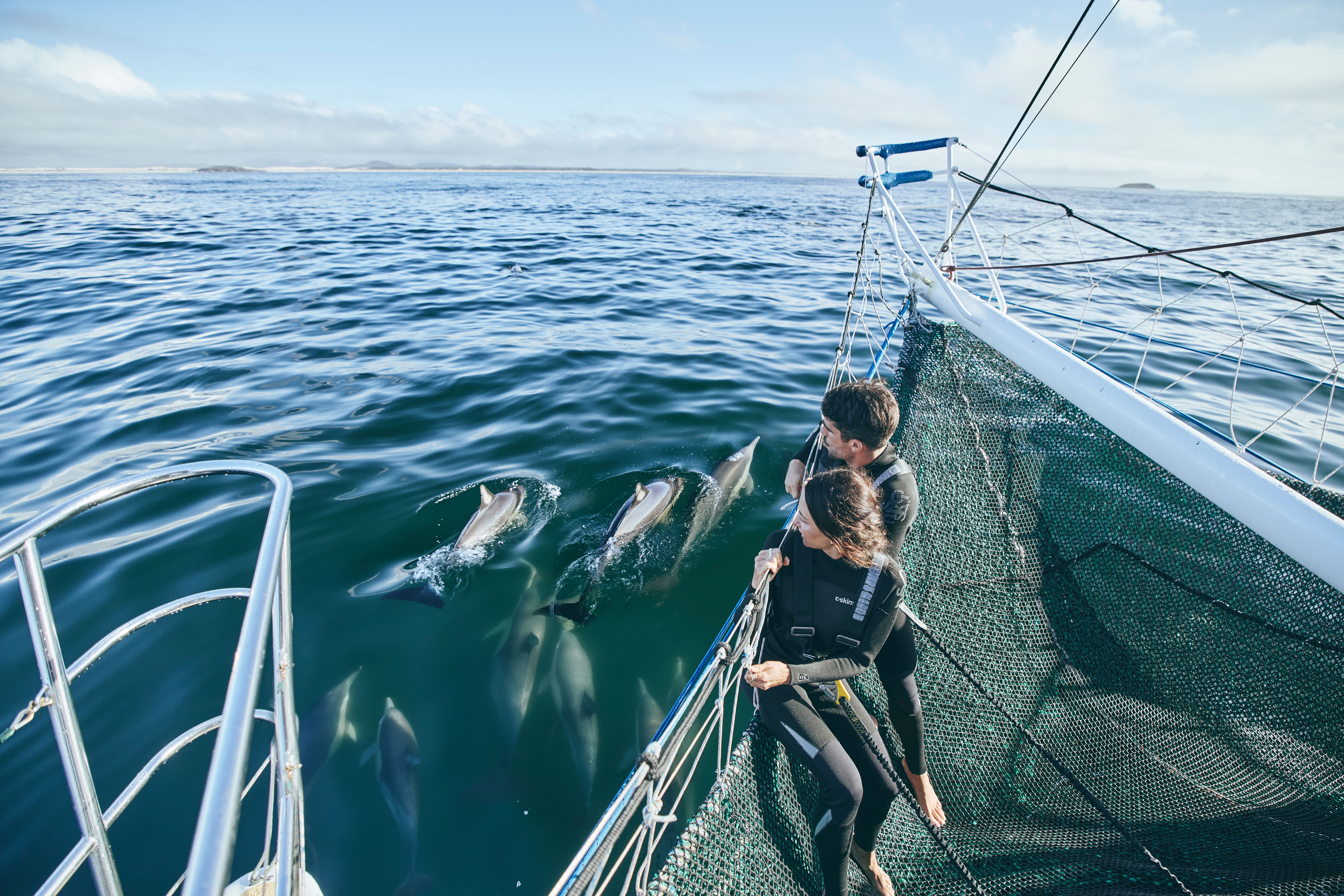Couple riding the boom net in their wetsuits with Dolphin Swim Australia watching dolphins playing in the waves at the front of the boat in Port Stephens