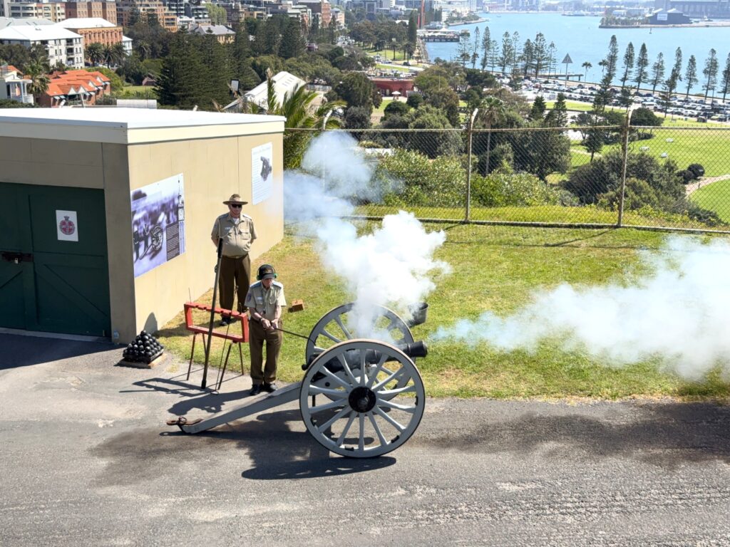 Firing of the canon at Fort Scratchley by volunteers in army uniform with Newcastle city in the background.